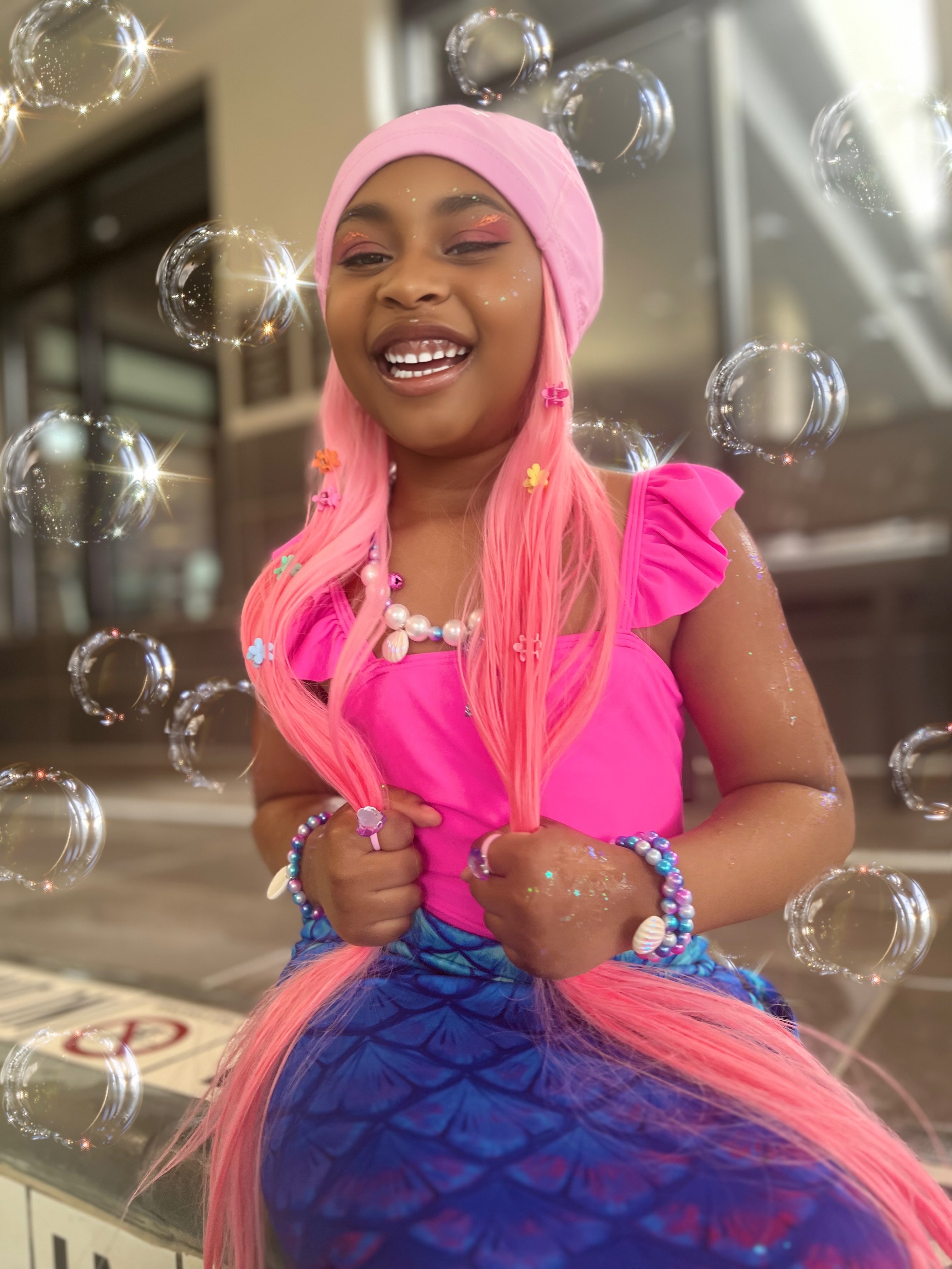 Girl in a pink mermaid cap and blue scale skirt, surrounded by soap bubbles.