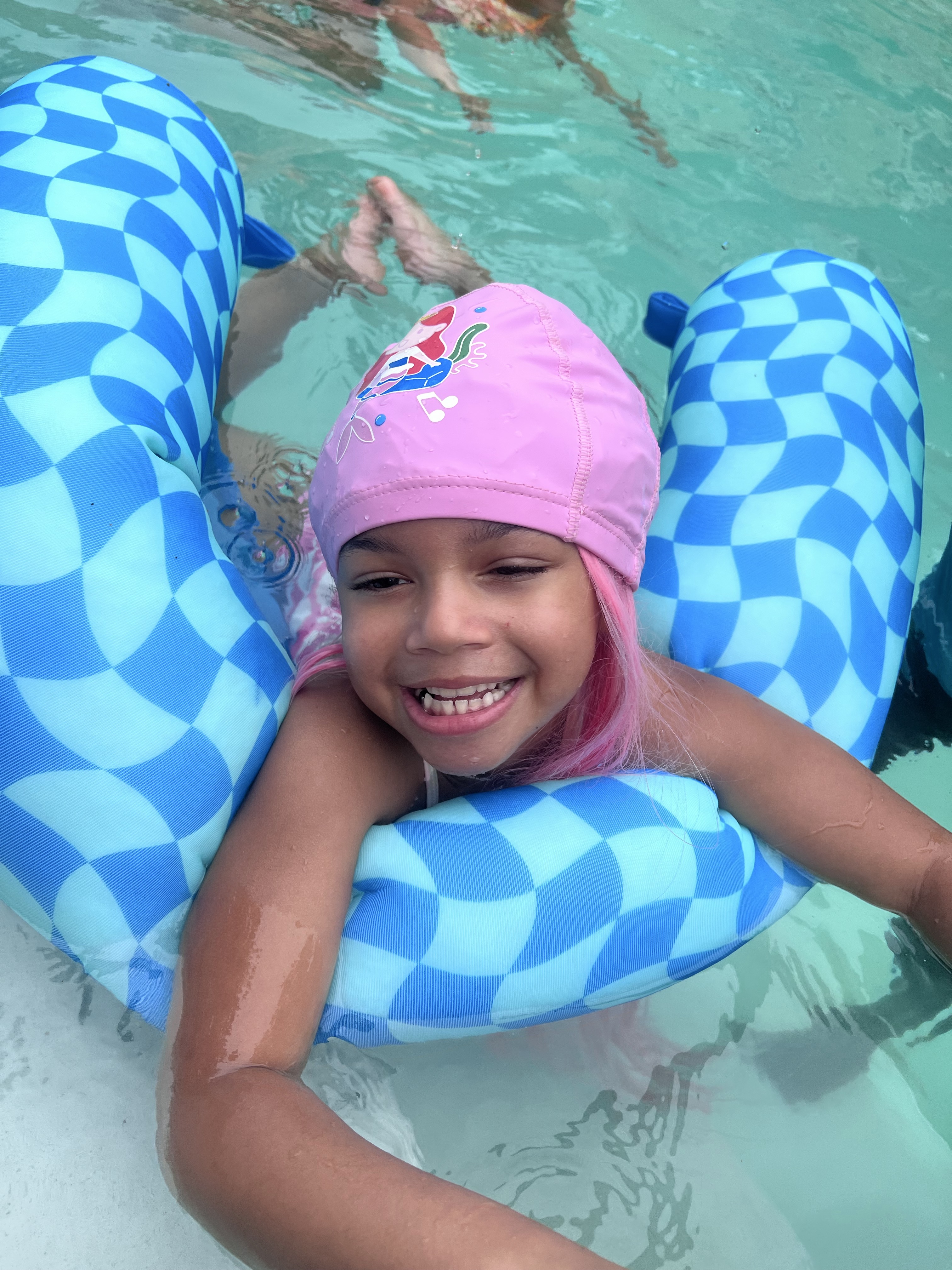 Child resting on a pool float wearing a pink cap with pink hair styled half up.