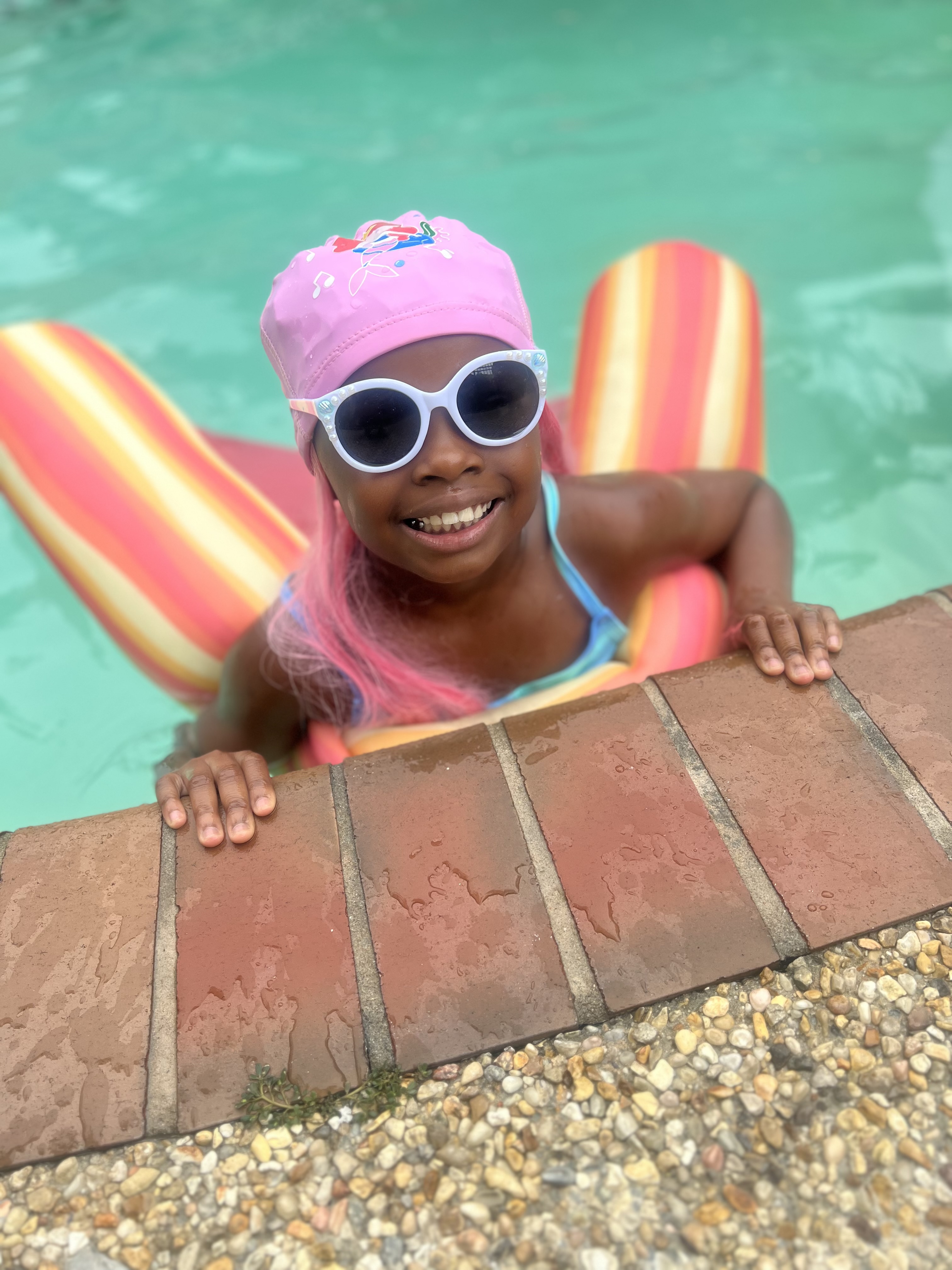 Child leaning on the pool edge wearing white round sunglasses and a pink mermaid cap.